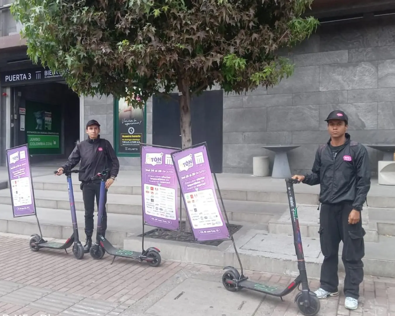 Dos promotores de Bagu Medios con scooters eléctricos y valla vertical frente a la entrada de una sede
