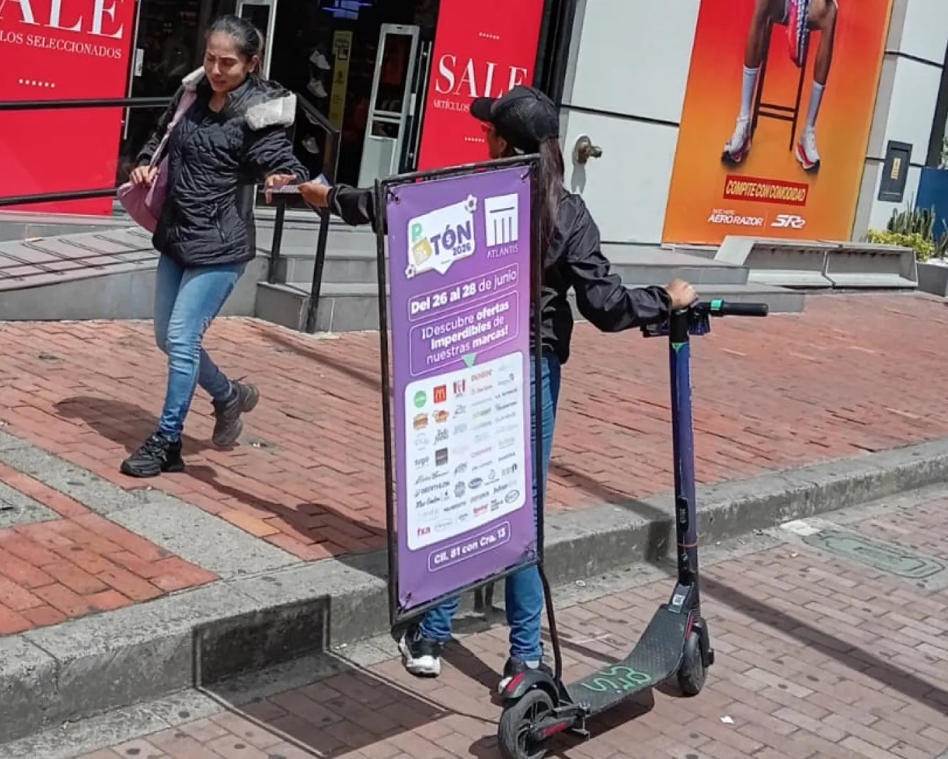 Promotoras de Bagu Medios con scooter valla de promoción comercial a la salida de un centro comercial